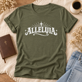 An army short-sleeve t-shirt with a white 'ALLELUIA' graphic printed on the chest, styled in a flat-lay arrangement with a brown leather journal, a cup of coffee, blue jeans, and dried flowers on a cream fabric background.