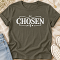 An army colored short-sleeve t-shirt with a white graphic print reading 'CHOSEN' in a distressed serif font, decorated with scrollwork and a small cross. The shirt is styled in a flat-lay arrangement on a light wood surface with a denim jacket, white sneakers, and small white flowers.