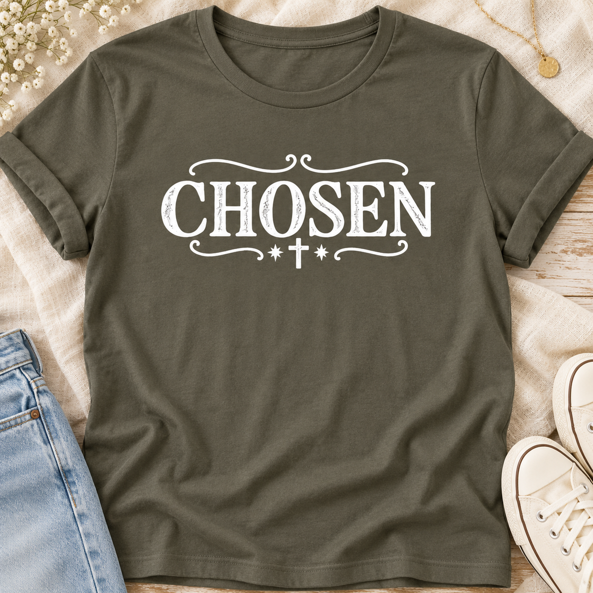 An army colored short-sleeve t-shirt with a white graphic print reading 'CHOSEN' in a distressed serif font, decorated with scrollwork and a small cross. The shirt is styled in a flat-lay arrangement on a light wood surface with a denim jacket, white sneakers, and small white flowers.