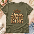 Army green Jesus Is King T-shirt with gold-toned lettering and a crown graphic, shown in a styled flat lay.