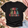 A black short-sleeve T-shirt laid flat with a colorful Christmas graphic reading 'Cozy Christmas' above three decorated trees. The shirt is styled on a cream knit blanket with holiday greenery, gold ornaments, a wrapped gift, pink ribbon, and denim shorts nearby.