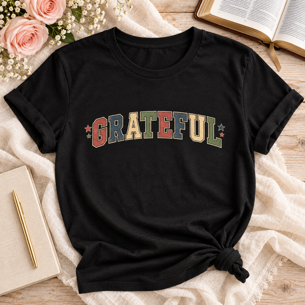 A black short-sleeve T-shirt laid flat on a light fabric surface, styled with pink roses, an open book, a notebook, and a pen. The shirt has the word "GRATEFUL" printed across the chest in multicolored block letters with small star accents.