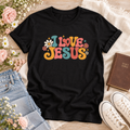 Black T-shirt reading “I Love Jesus” in colorful retro lettering, surrounded by daisies, small flowers, and sparkle accents.