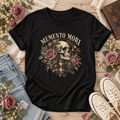 Black T-shirt with “Memento Mori” lettering above a vintage skull, mauve roses, leafy details, and an hourglass.