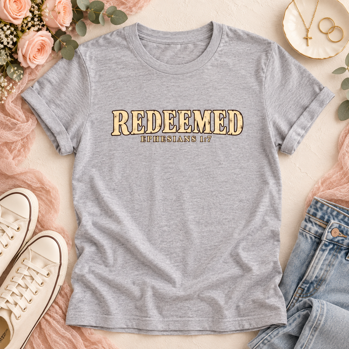 A heather gray short-sleeve t-shirt with 'REDEEMED' and 'EPHESIANS 1:7' printed on the chest, styled in a flat lay with jeans, white sneakers, pink fabric, flowers, and gold jewelry.