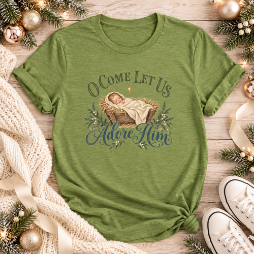 A green short-sleeve t-shirt with a Christmas-themed graphic reading 'O Come Let Us Adore Him' around an illustration of a baby in a manger.