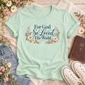 Mint T-shirt reading “For God So Loved the World” and “John 3:16,” with blue lettering framed by cream daisies, pink flowers, and leafy branches.