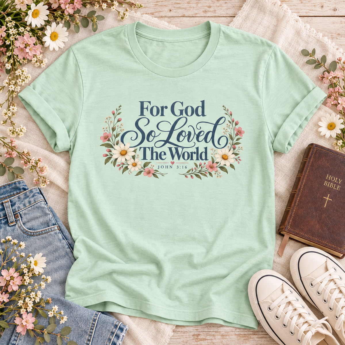 Mint T-shirt reading “For God So Loved the World” and “John 3:16,” with blue lettering framed by cream daisies, pink flowers, and leafy branches.