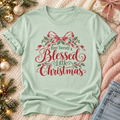 A mint green short-sleeve t-shirt with a red and green Christmas graphic reading 'Have Yourself a Blessed Little Christmas' surrounded by a red bow and holly leaves, styled in a flat lay on a pink knit blanket with holiday decorations and jeans.