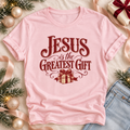 A light pink short-sleeve t-shirt with a maroon graphic reading 'JESUS is the GREATEST GIFT' above a wrapped gift illustration.