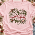 A pink short-sleeve crewneck t-shirt featuring a festive graphic print that reads 'Let Heaven and Nature Sing' in dark green and red cursive script, decorated with pine branches, red berries, and a small bird. 