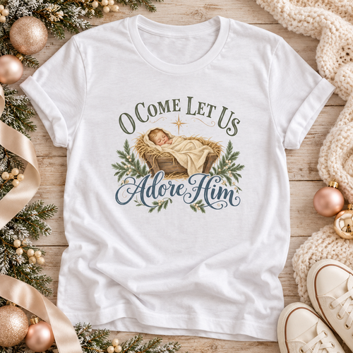 A white short-sleeve graphic T-shirt features a printed illustration of a baby in a manger with the text 'O COME LET US Adore HIM' in green and blue lettering.